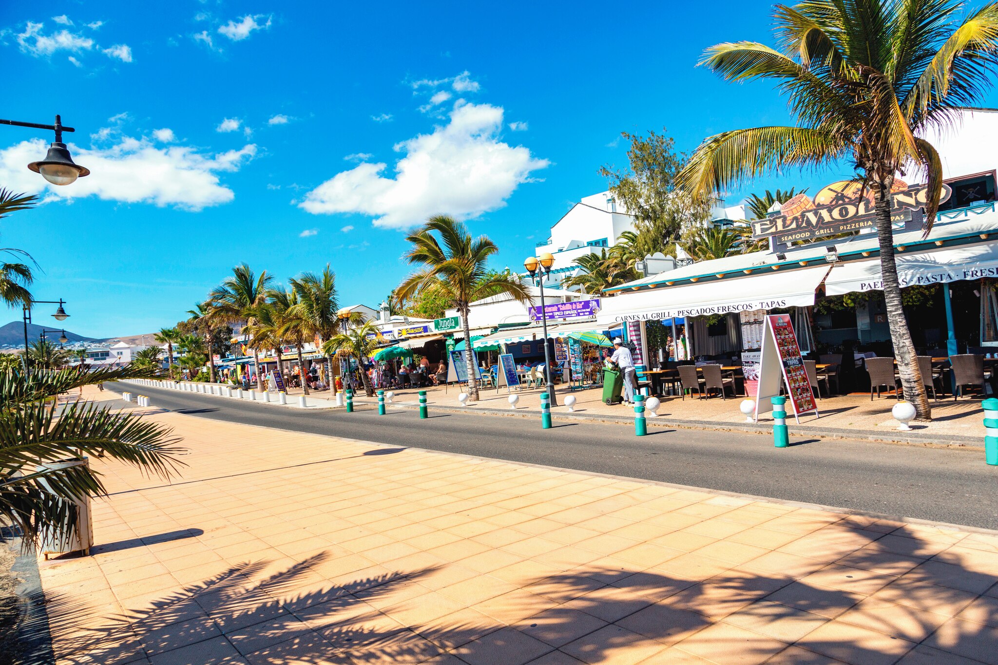 Colorful Los Pocillos promenade lined with palm trees, restaurants, and shops under a bright blue sky.