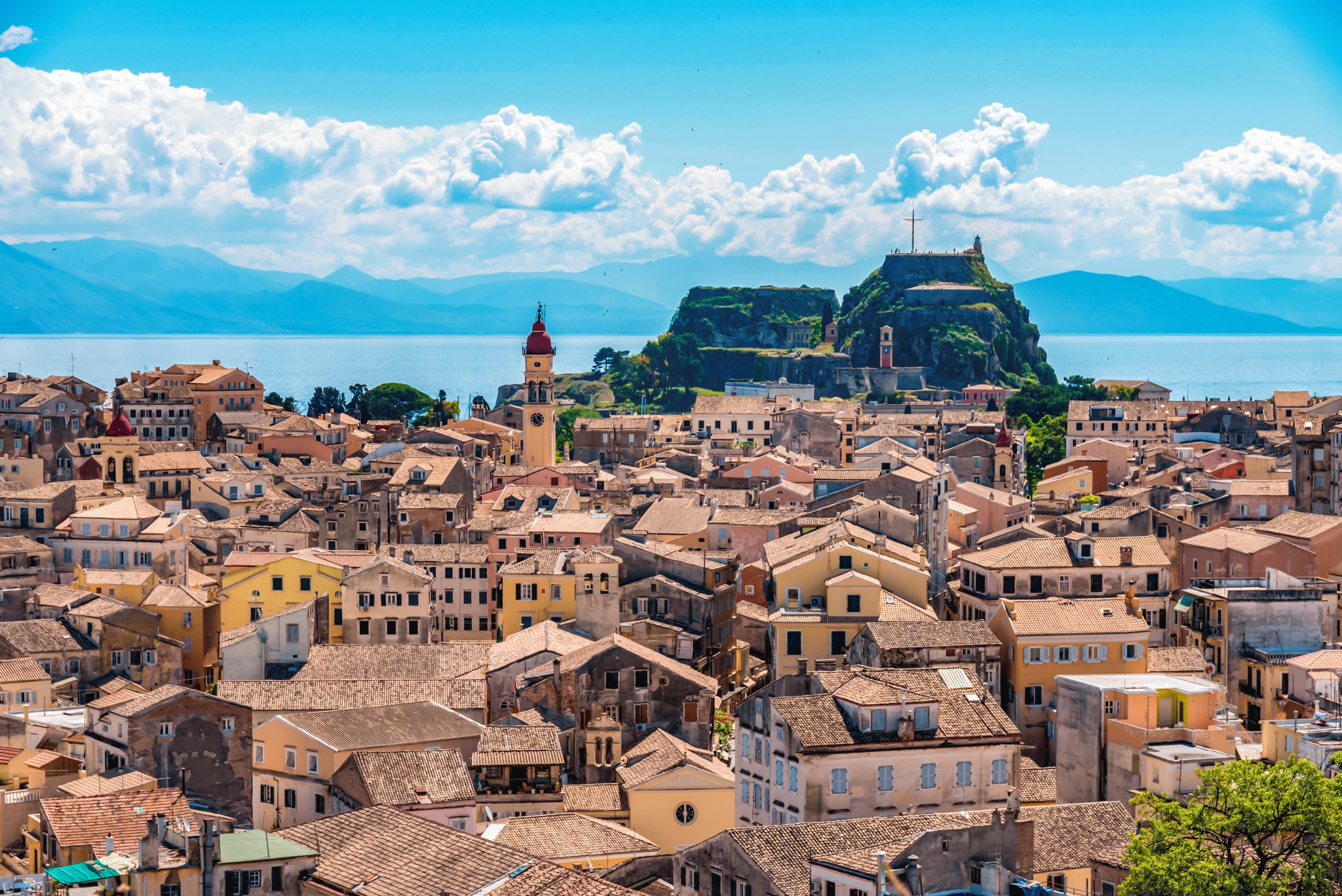 A panoramic view of Corfu Old Town, showcasing colorful buildings, a lighthouse, and a mountainous backdrop against the shimmering sea.