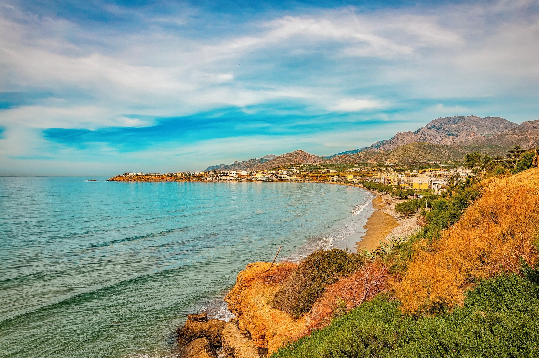 Scenic view of Makry Gialos, a coastal village in Crete, with a sandy beach, clear waters, and surrounding mountains.