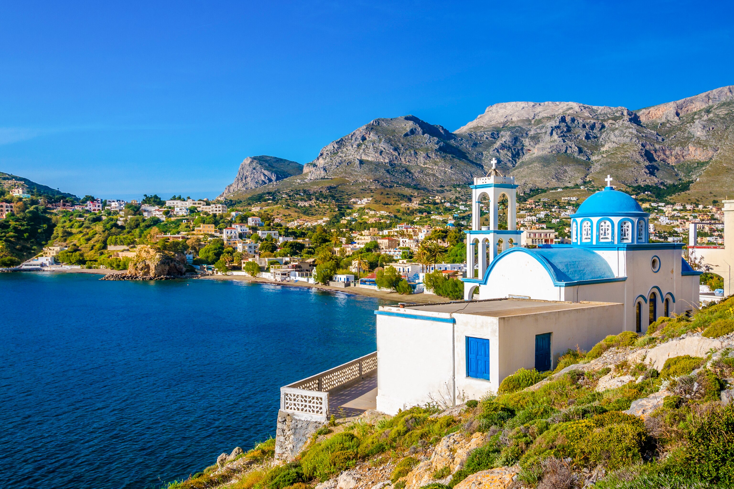 Coastal view of Panormos on Kalymnos Island, featuring a blue-domed church and vibrant greenery against a mountainous backdrop.
