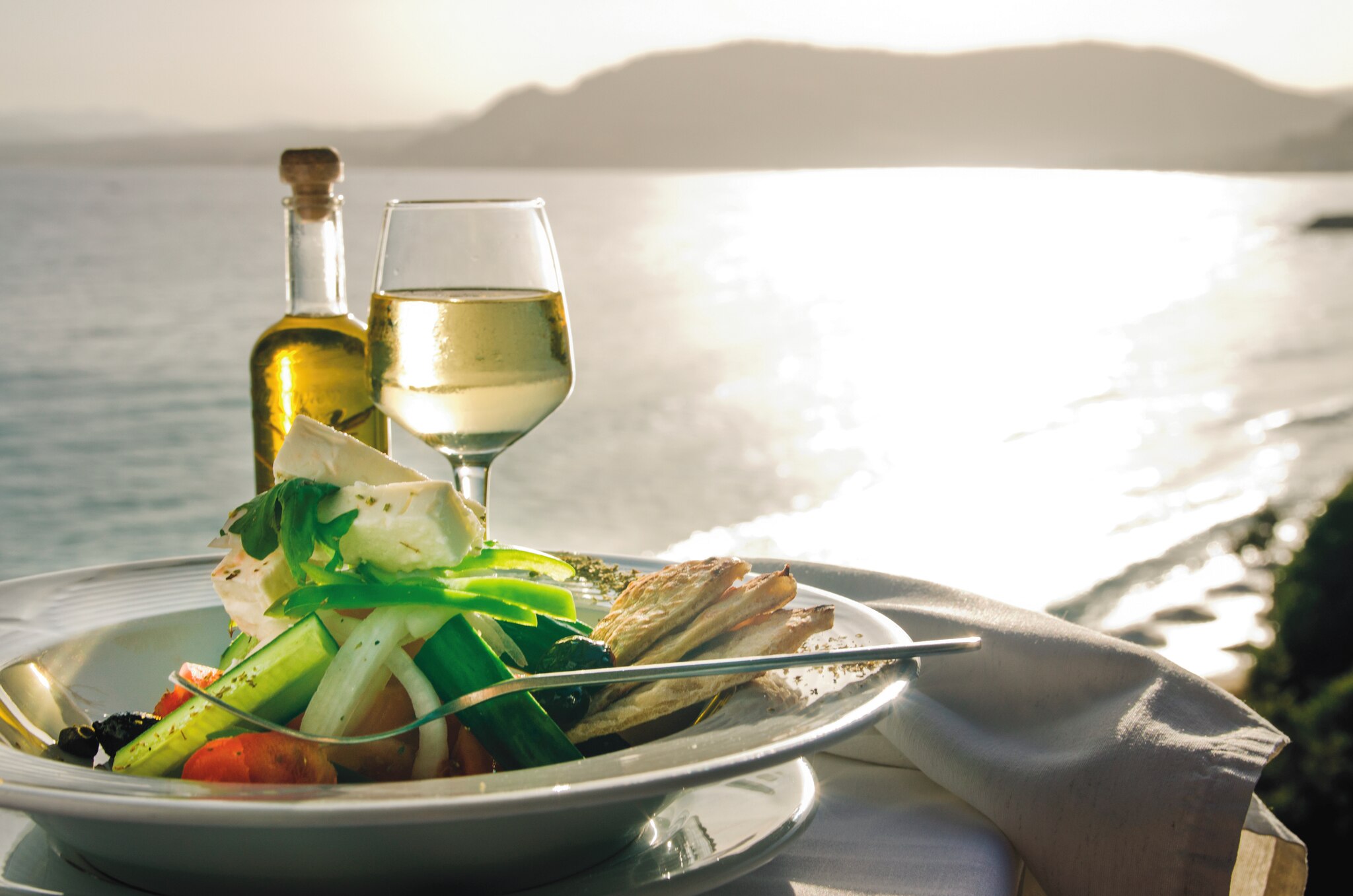 A plate of seafood and feta salad, accompanied by a glass of white wine and a bottle of olive oil, set against a shimmering sea backdrop in Pastida.