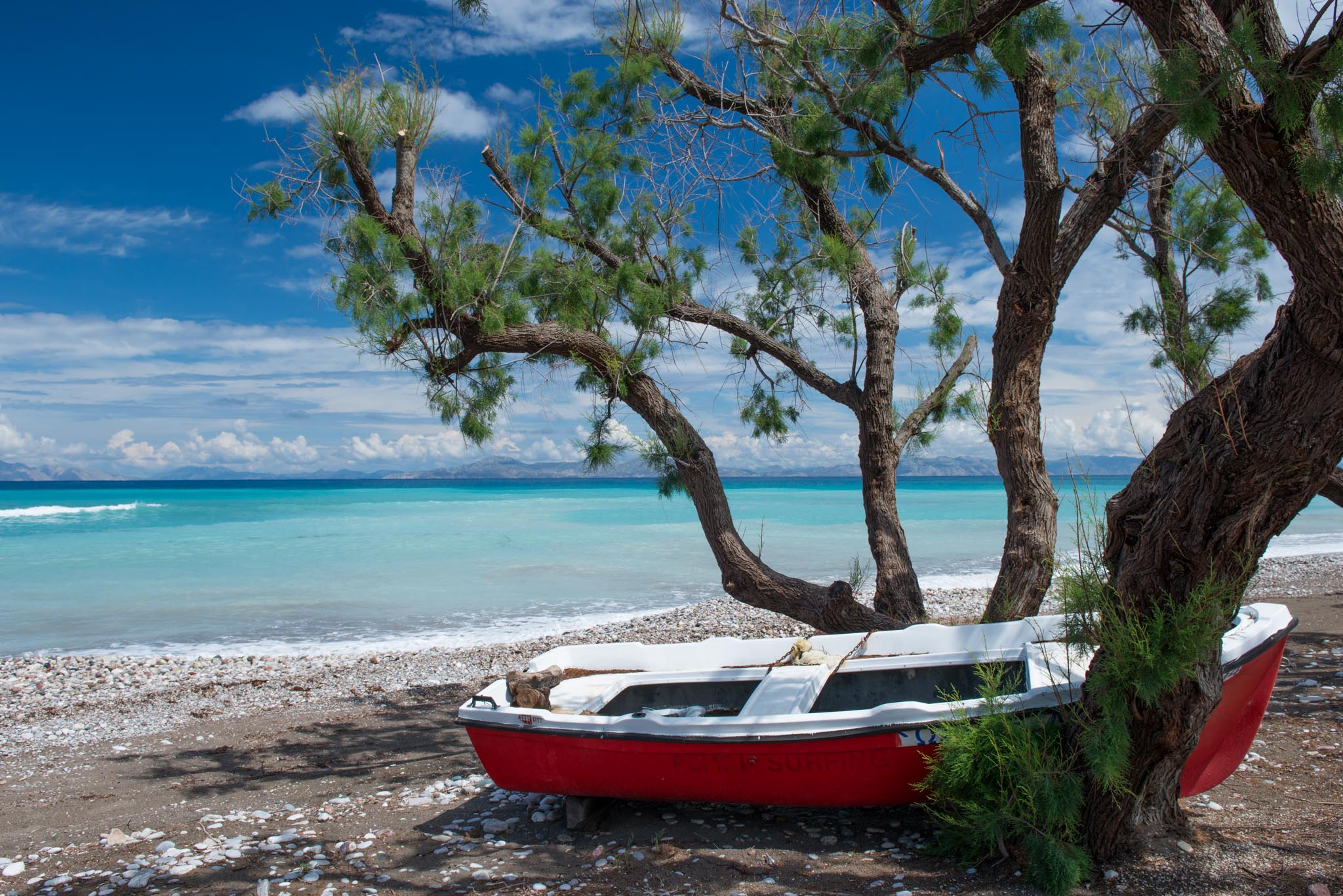A tranquil beach scene at Theologos, featuring a tree by the shore and turquoise waters under a blue sky with fluffy clouds.