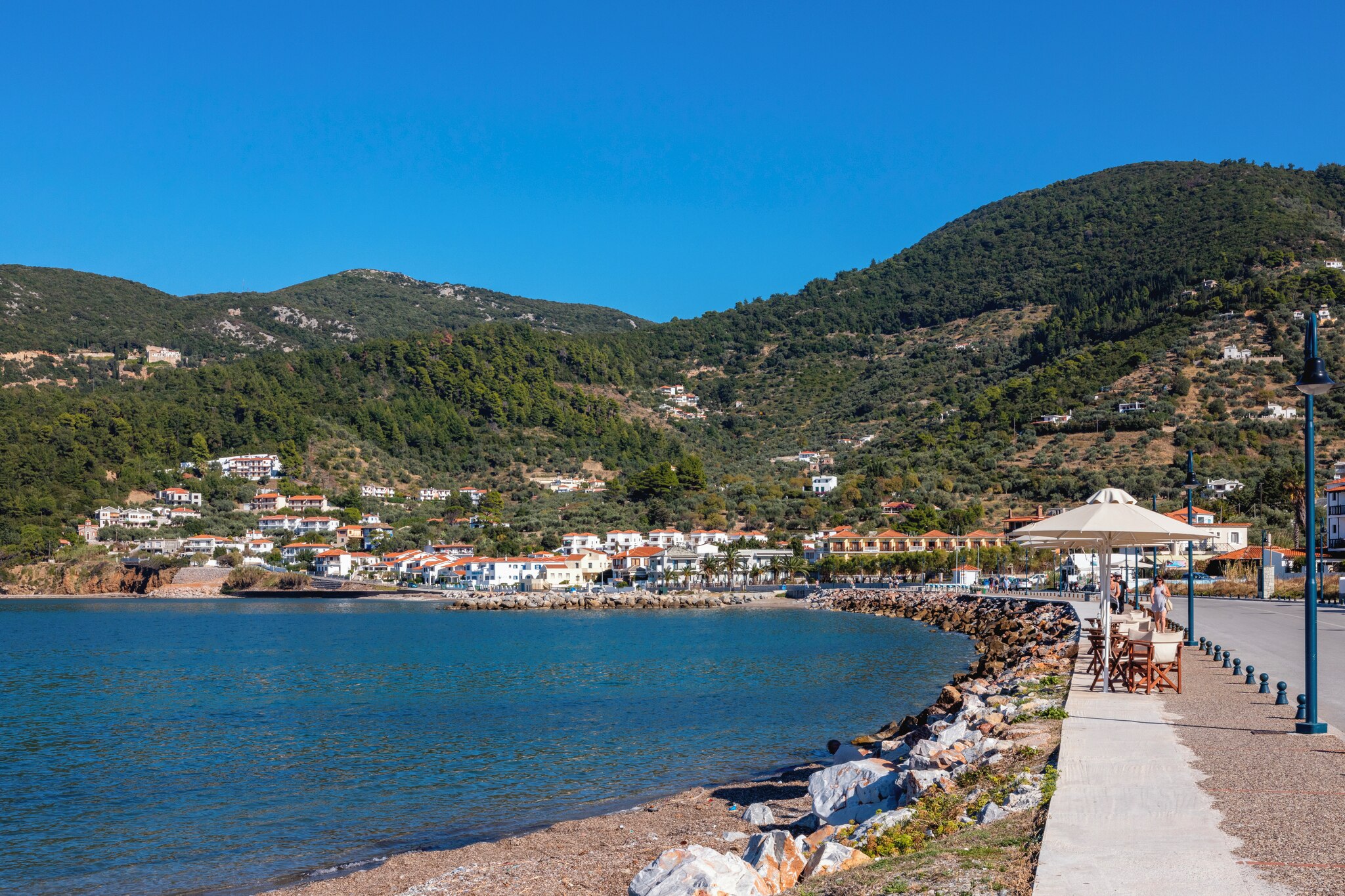 Charming coastal view of Skopelos Island featuring white villas, green hills, and a sunny seaside promenade with visitors.