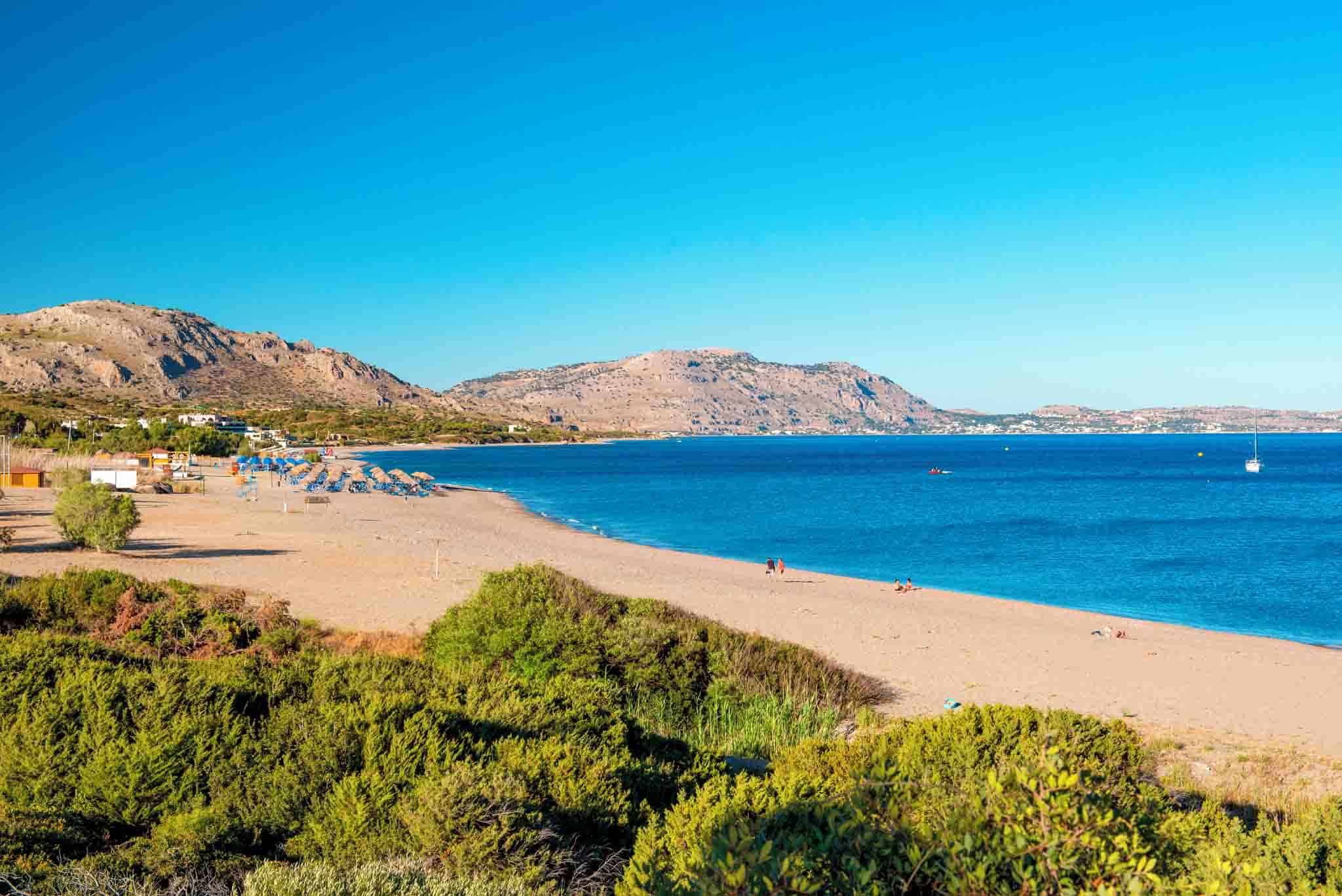 Kiotari beach in Rhodes, with sandy shores, straw umbrellas, and a turquoise sea against rugged hills in the background.