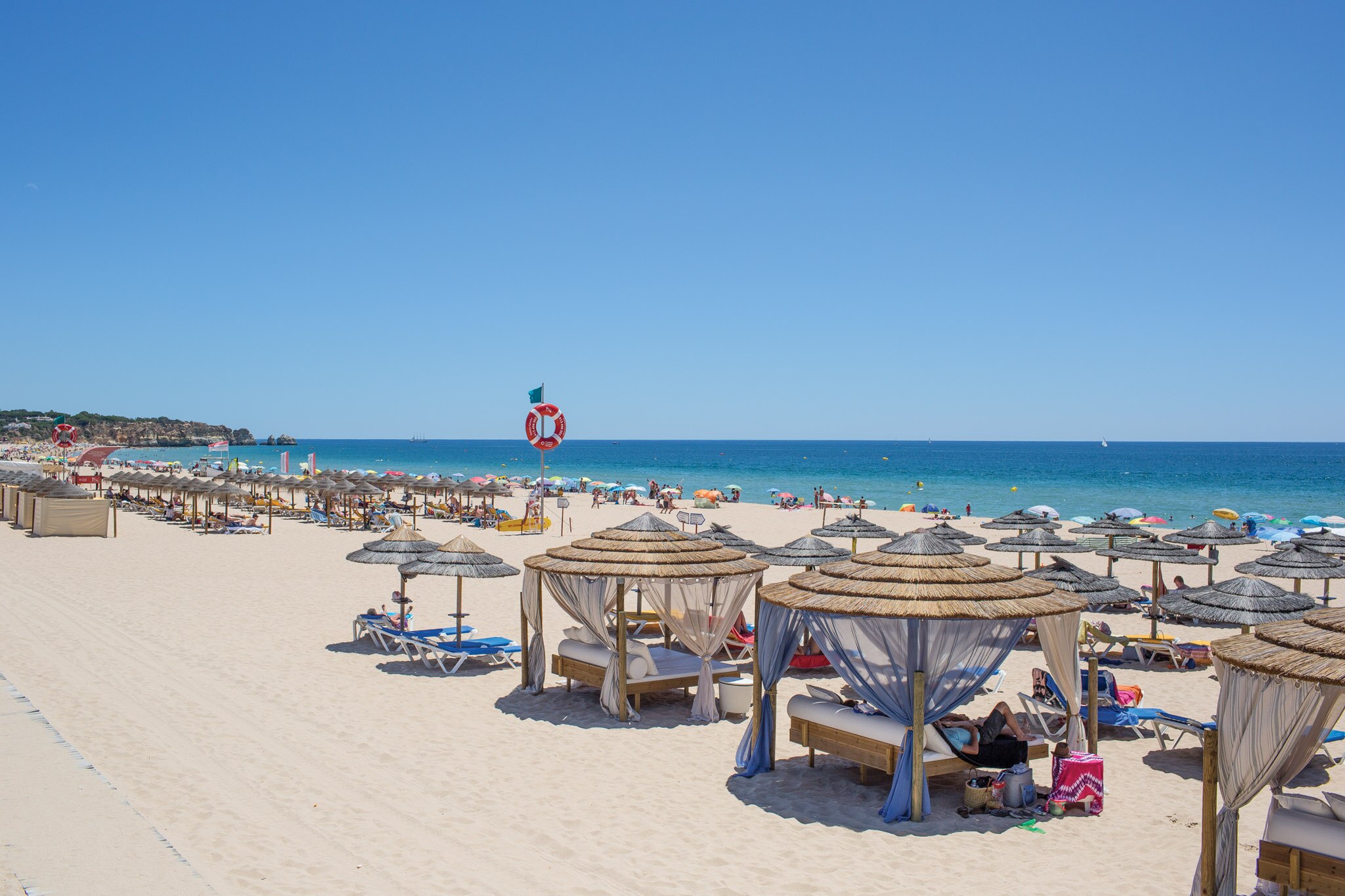A sunny day at Praia de Alvor beach, featuring straw umbrellas, sunbathers, and vibrant waters under a clear blue sky.