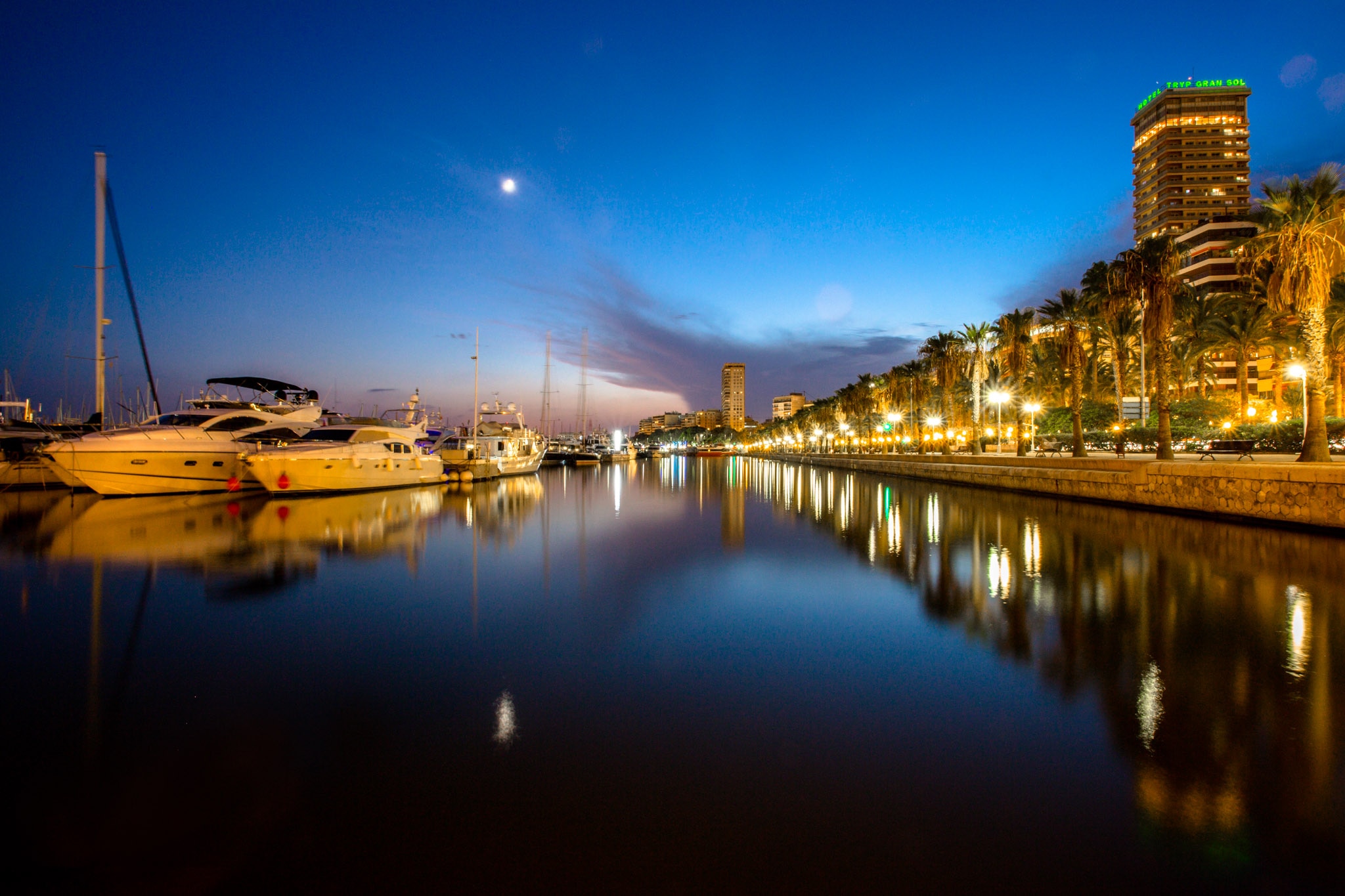 Alicante Marina at dusk, featuring lined yachts and palm trees reflecting in still waters under a colorful sky.