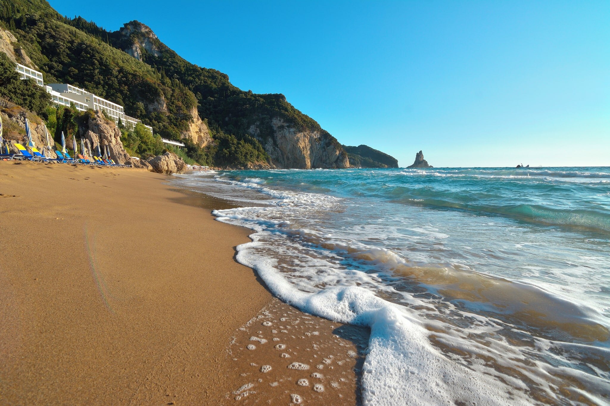 Scenic view of Aghios Gordios beach, featuring golden sand, colorful loungers, and tranquil waves lapping at the shore.