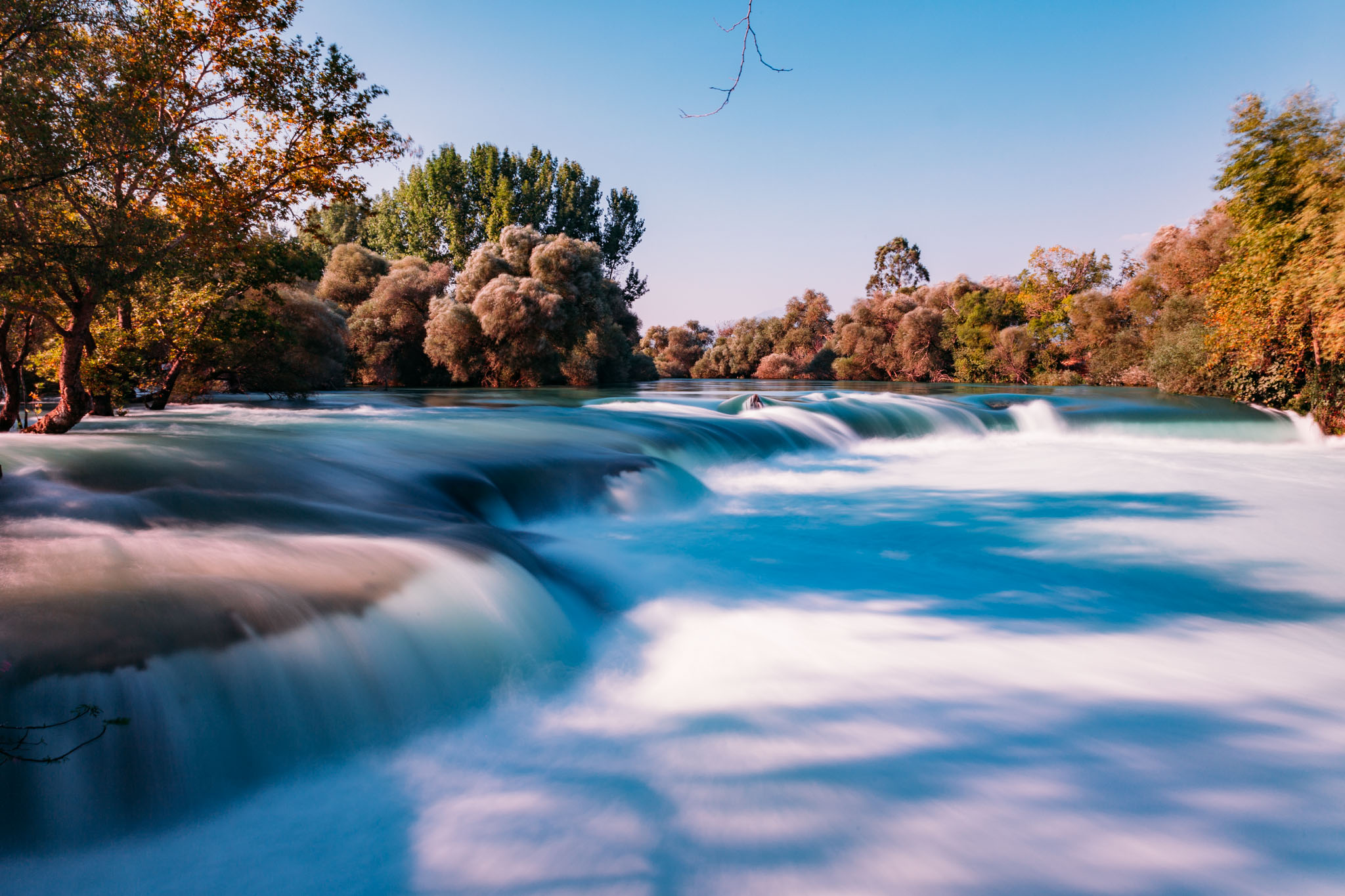 A serene view of Manavgat Waterfall with flowing turquoise waters and lush greenery in the background under a clear sky.