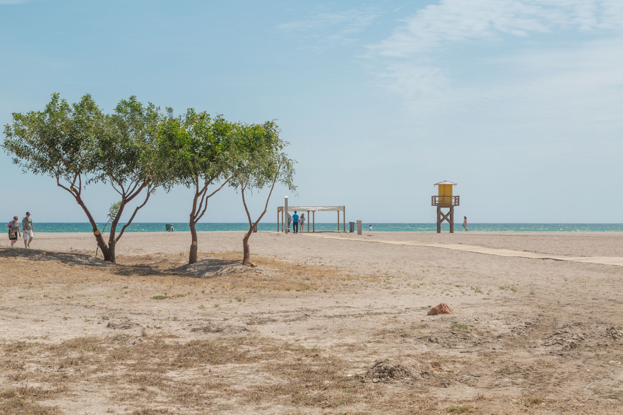 Beach scene at Playa el Playazo with trees, a lifeguard tower, and people walking along the sandy shore under a blue sky.