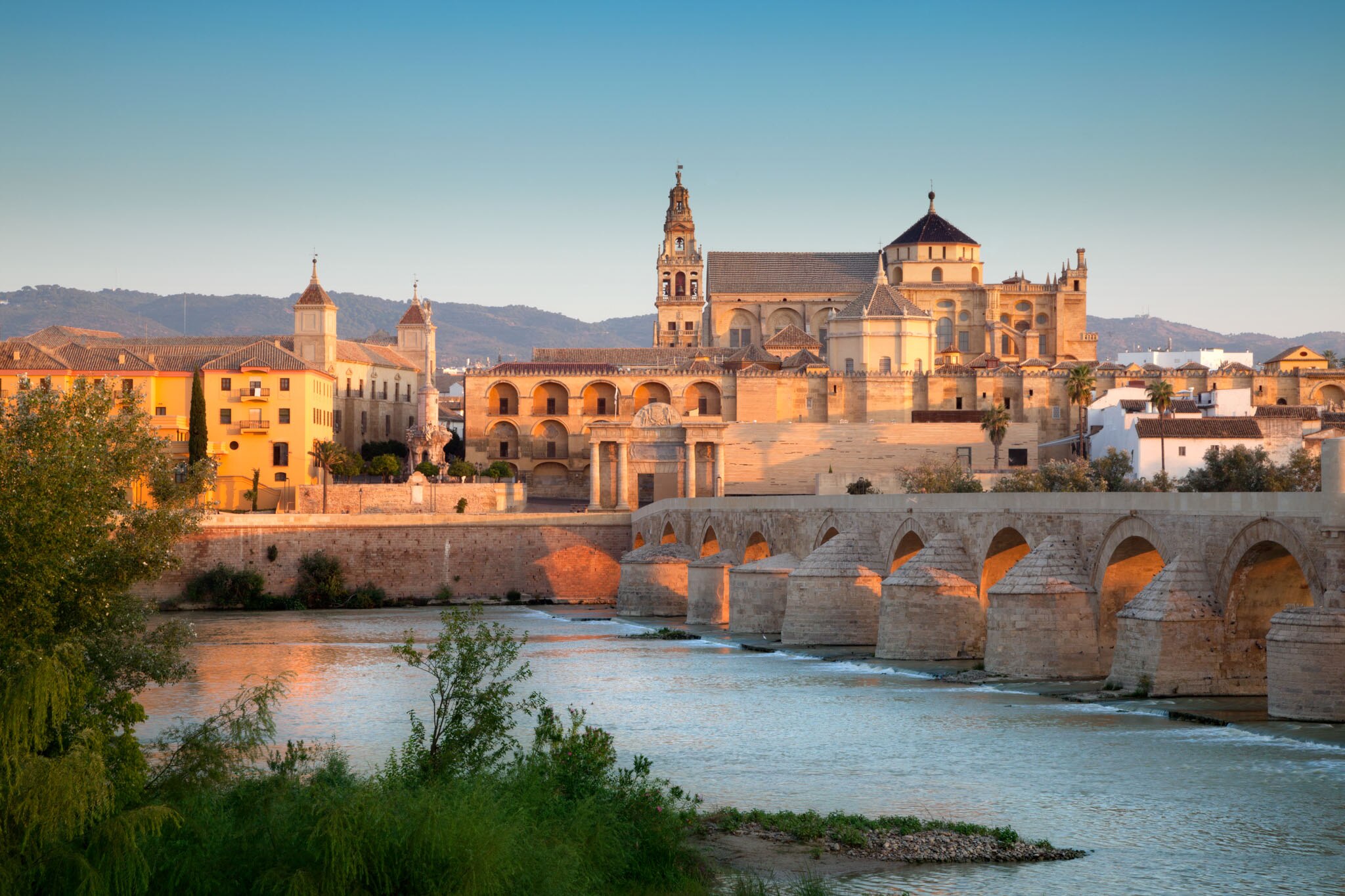 Scenic view of Cordoba, showcasing historic architecture and the Roman bridge against a backdrop of mountains and sunrise.
