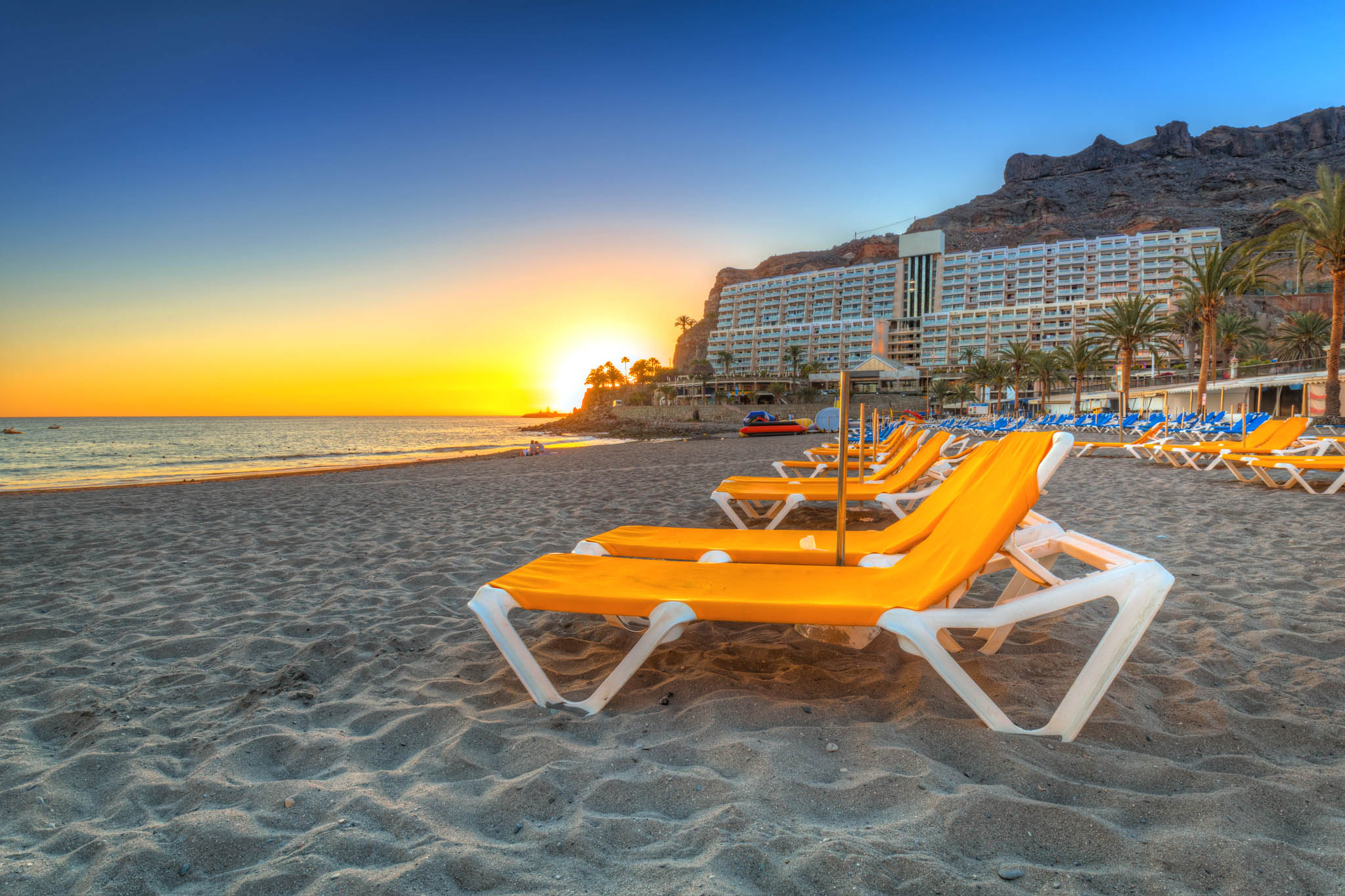 Golden sunset over Playa Taurito, with empty orange beach loungers on sandy shore and palm trees in the background.