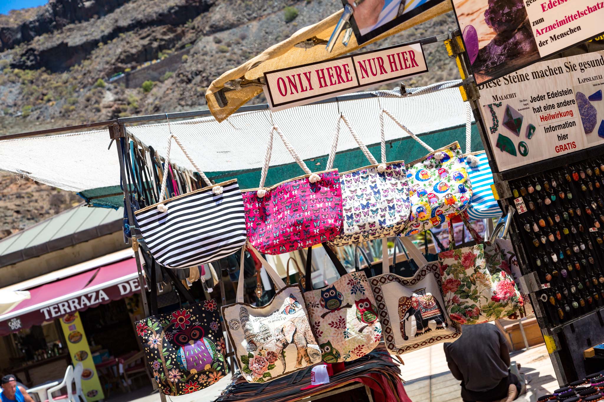 Vibrant bags displayed for sale at Mogan Market, with various patterns against a sunny backdrop and a vendor stall nearby.