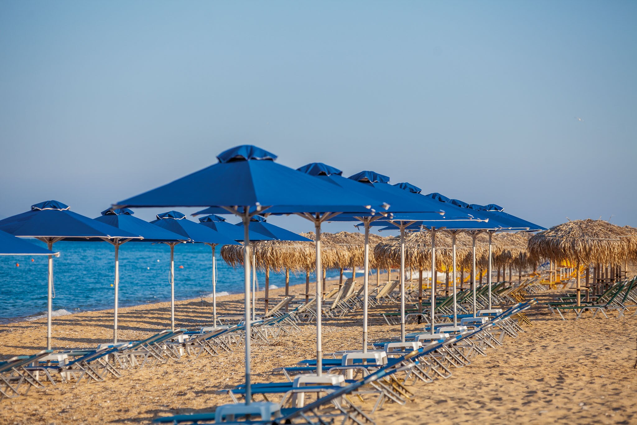 Blue beach umbrellas lined up at Skala Beach, overlooking the calm sea and sandy shore under a clear sky.