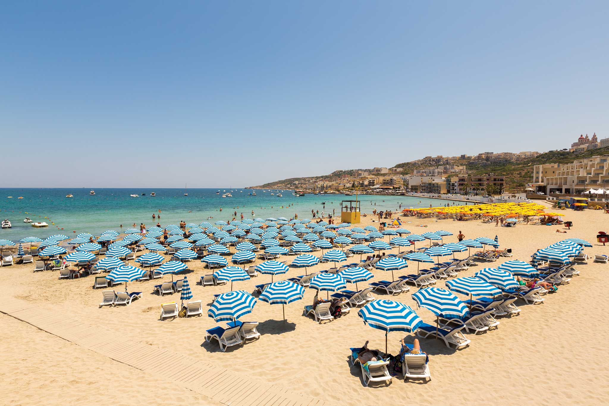 Mellieha Bay Beach features sunbathers under blue-striped umbrellas, clear waters, and a charming coastal backdrop in Malta.