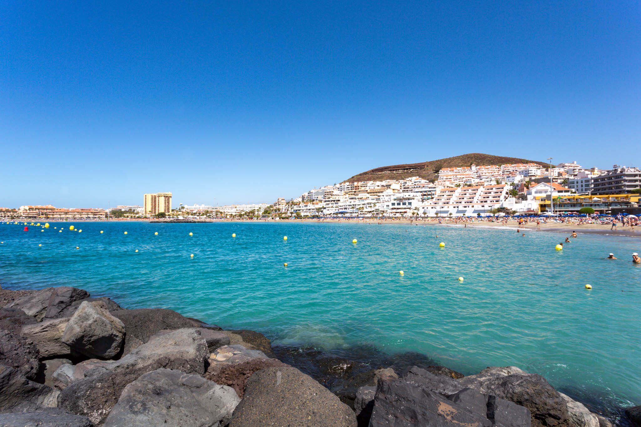 View of Playa de las Vistas, featuring clear blue water, sandy beach, and white buildings under a bright blue sky.
