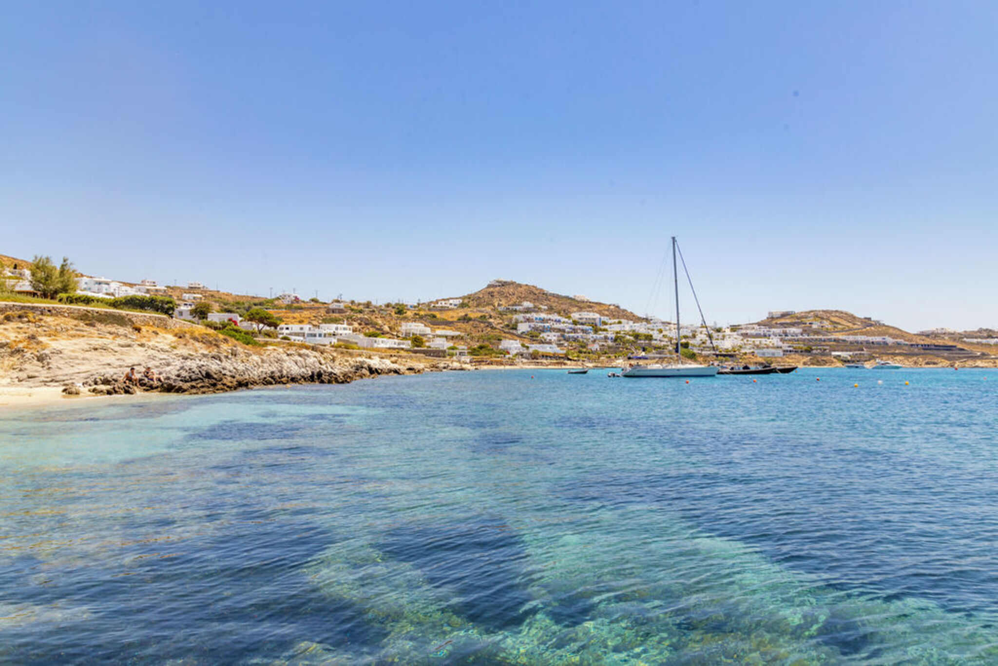 A Agios Ioannis beach, featuring rocky shores, crystal-clear waters, and picturesque white-washed buildings on a hillside.
