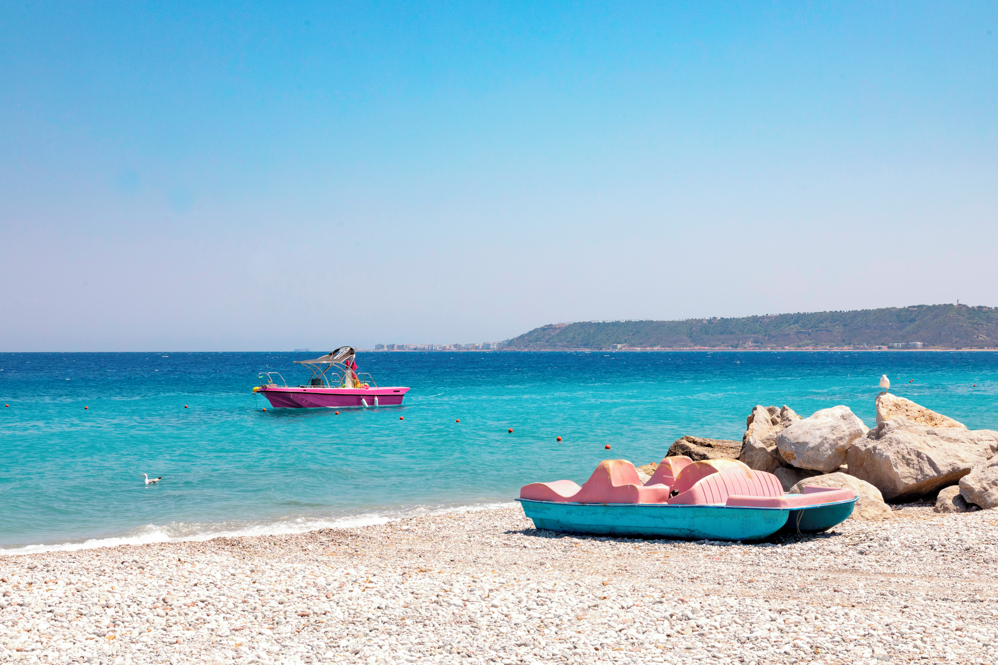 A pink boat floats in the turquoise sea of Ialyssos near a rocky shore, with a green hill in the background.