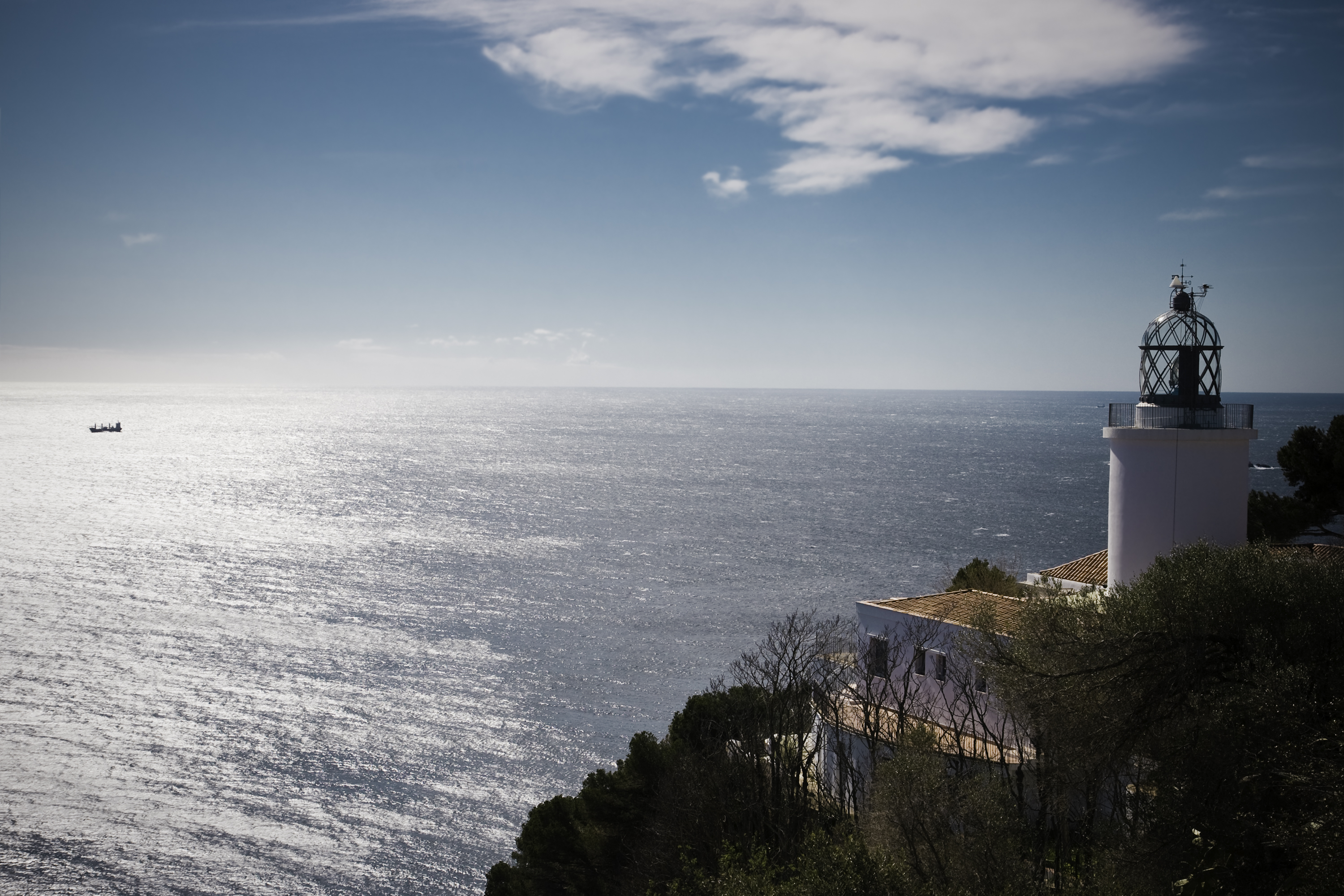 Sant Sebastià Lighthouse stands near the coast, overlooking a tranquil sea, with sunlight shimmering on the water's surface.