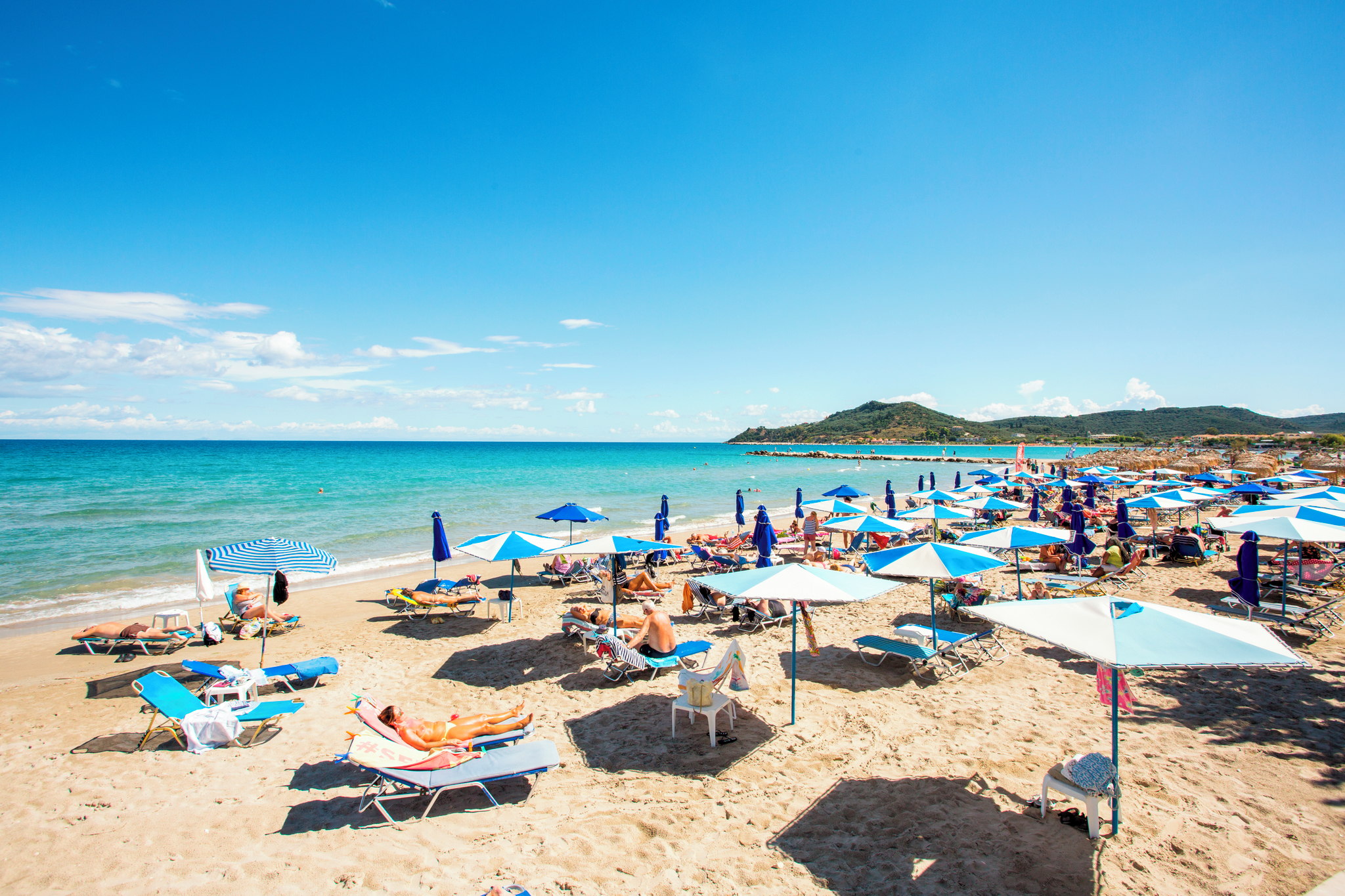 A sunny beach at Alykes, with sunbathers under blue and white umbrellas, clear blue sea and green hills in the background.