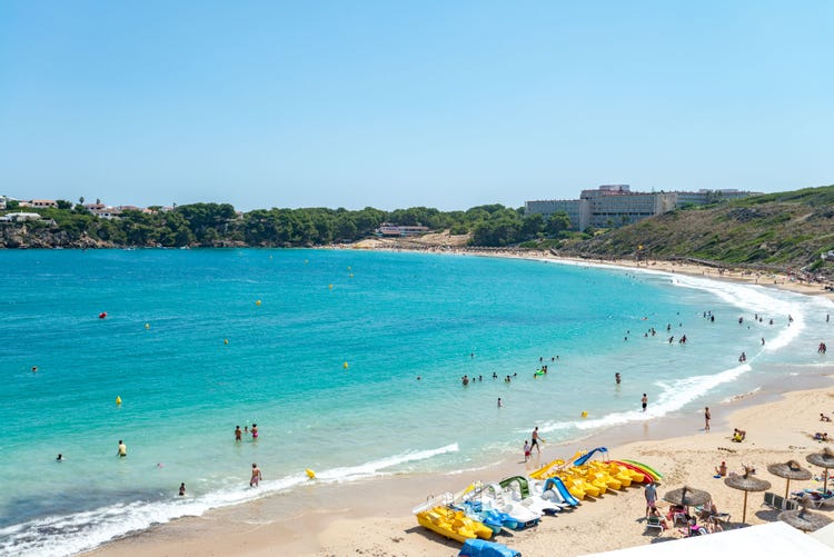 A scenic beach view of Arenal D'en Castell, with turquoise waters, sandy shore, and people enjoying the sun and sea.
