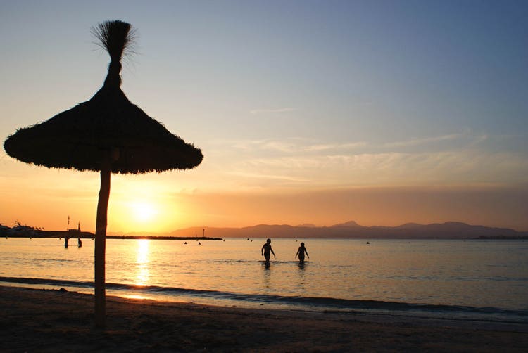 A serene sunset at El Arenal beach, featuring a silhouette of a thatched umbrella with vibrant orange skies in the background.