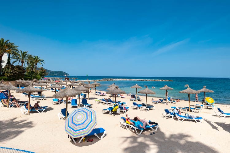 Sun-soaked Cala Bona beach with straw umbrellas, sun loungers, and clear waters under a bright sky, filled with beachgoers.