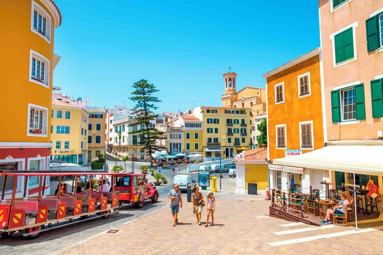 A Mahon square featuring colorful buildings, outdoor seating, and sunny blue skies, evoking a lively Mediterranean atmosphere.