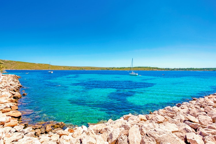 Sunny view of Playas de Fornells with clear turquoise waters, rocky shoreline, and boats on a serene Mediterranean day.
