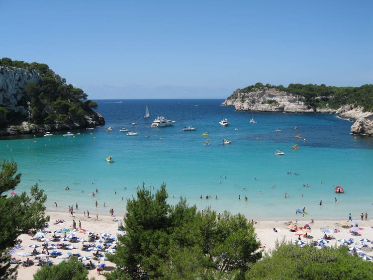 A picturesque view of Cala Galdana, featuring turquoise calm waters, boats, and rocky cliffs under a clear blue sky.