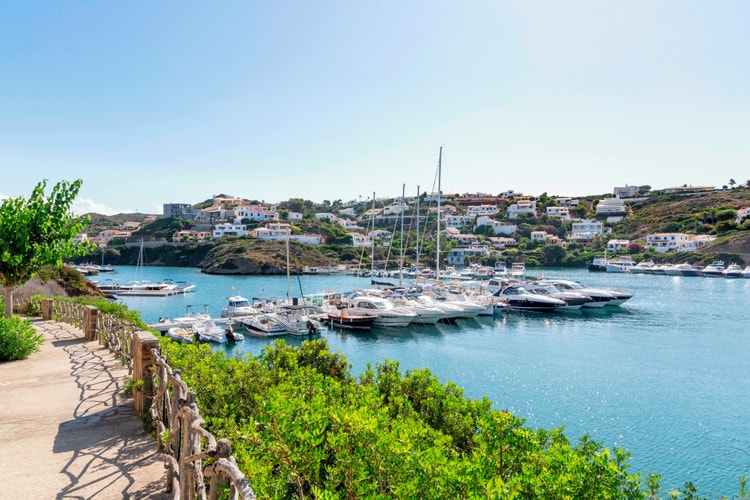Coastal view of Cala Llonga, Menorca, featuring sailboats in a serene bay and picturesque hillside homes.