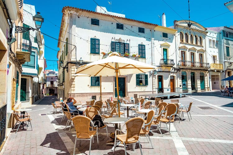 Sunny street view of Alaior with outdoor café seating and colorful buildings in a charming, vibrant atmosphere.