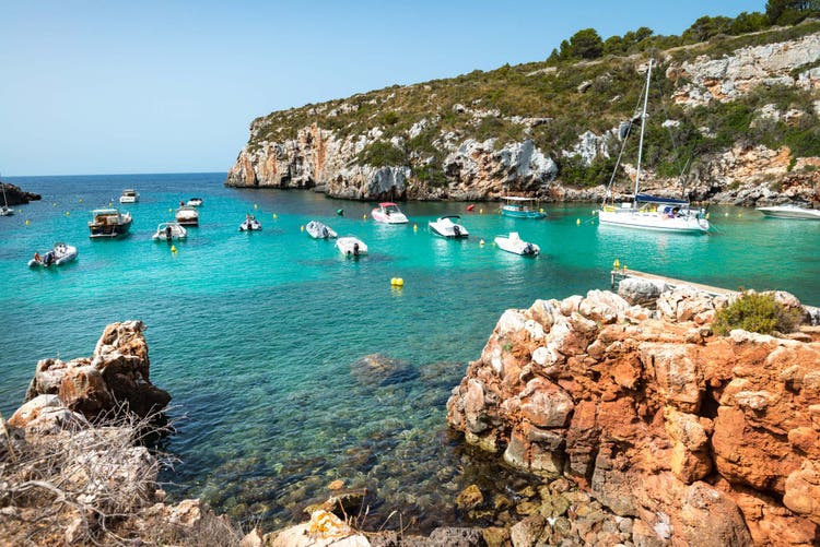 Serene Cala Canutells with turquoise waters, dotted with white boats and rocky shores, inviting a peaceful sea escape.