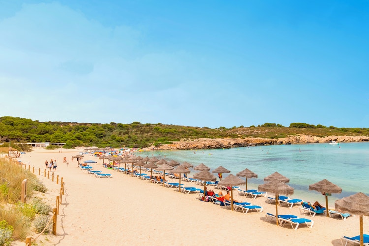 A busy beach at Son Parc filled with people enjoying the sun and a few boats floating in the water.