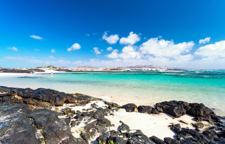 A scenic view of El Cotillo beach with turquoise waters, sandy shores, and mountain landscape on the background.