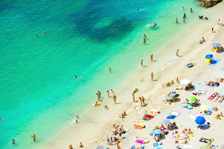 Argineguín beach with people swimming in turquoise water and sunbathing on the sandy shore under bright umbrellas.