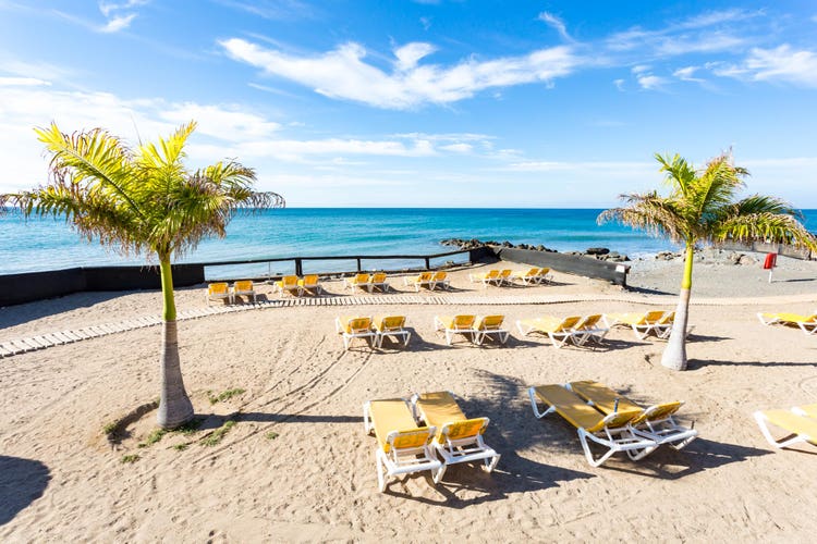 Sunny beach scene at Bahia Feliz with yellow lounge chairs and palm trees, overlooking calm blue waters and a clear sky.