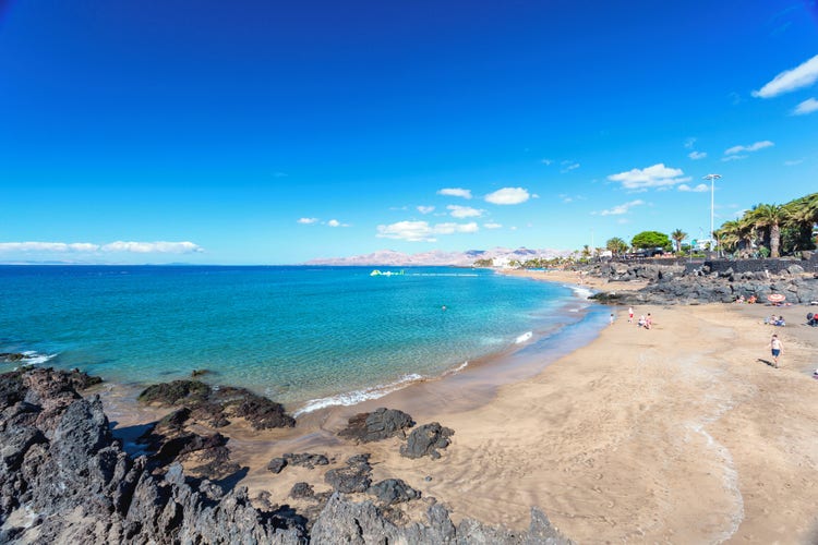 A picturesque beach in the Puerto del Carmen, Lanzarote, featuring golden sands and clear blue waters under a bright sky.