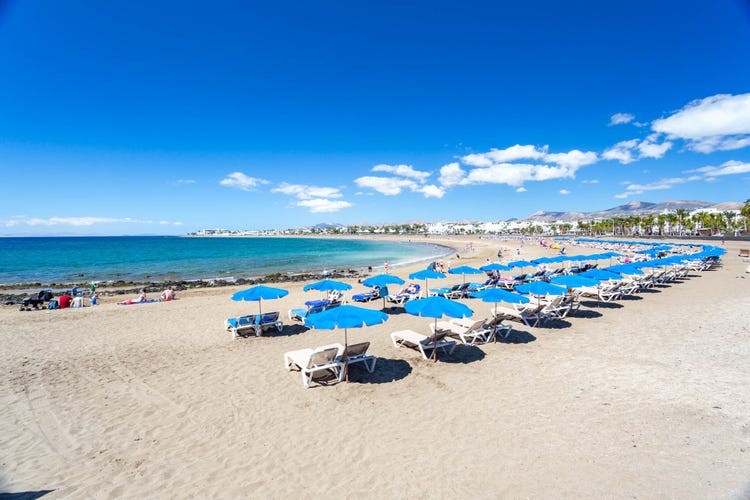 Playa De Los Pocillos beach featuring blue umbrellas, sun loungers, sunbathers, and clear blue sea.