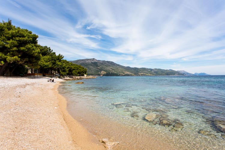 A beach in Orebić, Croatia, with turquoise waters, sandy shore, and green pine trees, against rugged mountains in the background.