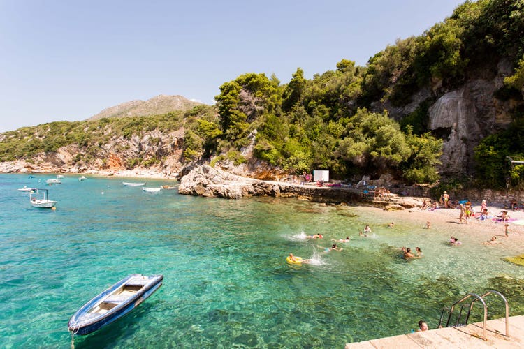 A sunny beach in Orasac, featuring clear turquoise water, boats, and people enjoying the seaside among lush green hills.