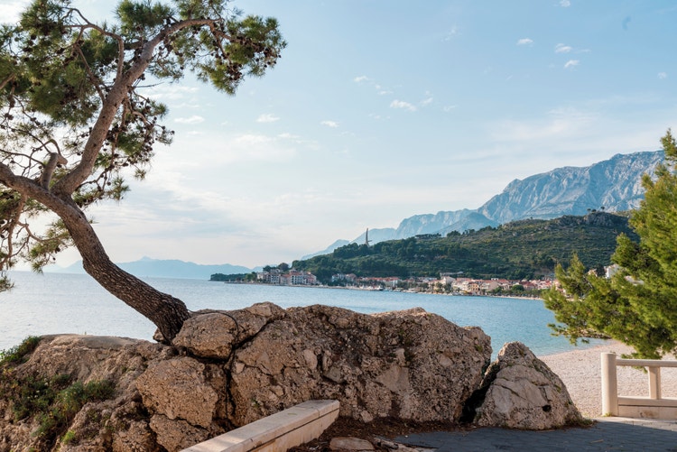 Scenic view of Podgora, Croatia, featuring a coastline with colorful buildings, mountains, and a clear blue sky.
