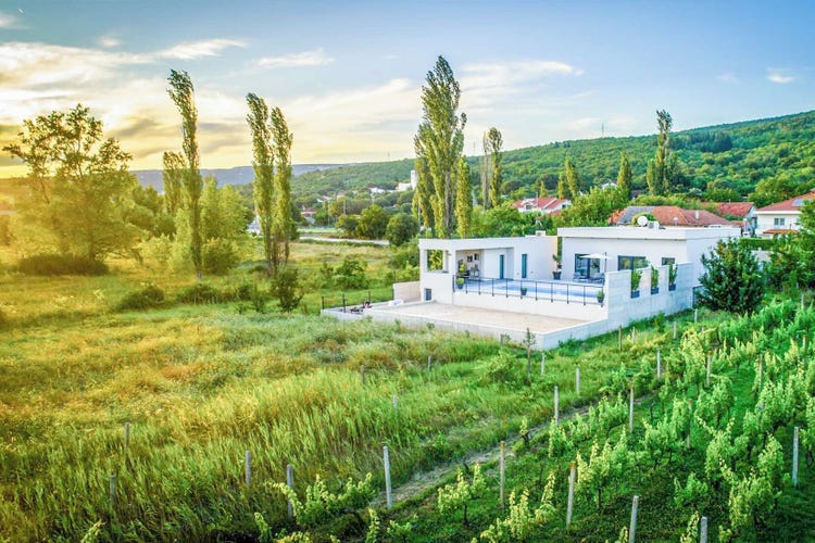 A modern white house with a large terrace, surrounded by lush greenery and mountains in Imotski, Croatia, during sunset.