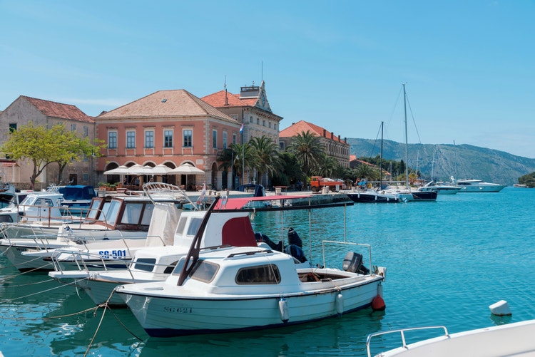 A coastal scene in Stari Grad, featuring colorful boats, palm trees, and historic buildings along the turquoise harbor.