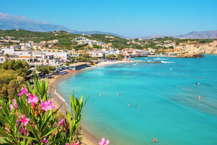 Aerial view of Almyrida beach, featuring turquoise waters, sandy shores, vibrant flowers, and a vibrant Mediterranean town.