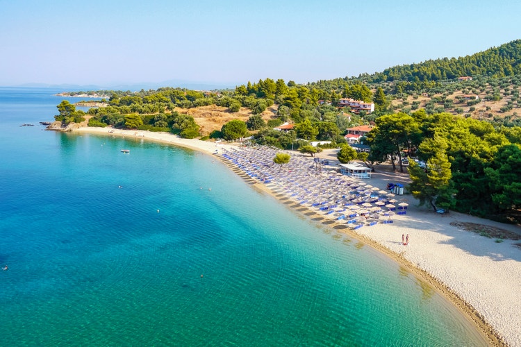 The beach of Lagomandra with its turquoise waters, sun loungers with blue parasols and the lush greenery in the background.