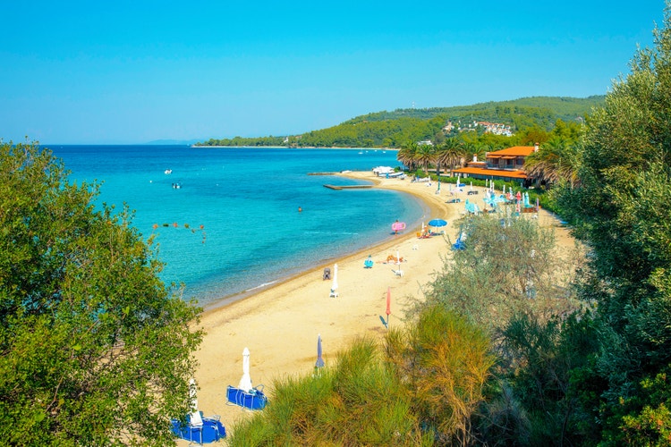 Akti Elias beach with golden sand, turquoise waters, beach umbrellas, and palm trees, set against a sunny coastal backdrop.