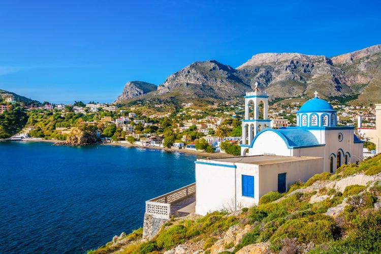 Coastal view of Panormos on Kalymnos Island, featuring a blue-domed church and vibrant greenery against a mountainous backdrop.