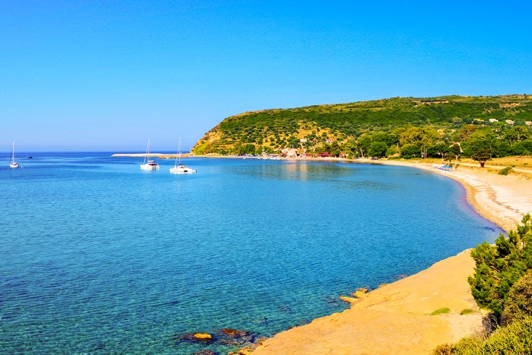 A serene view of Katelios beach, featuring calm turquoise waters, sailboats, and lush green hills in the background.
