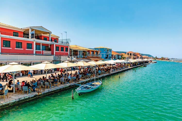 Outdoor cafes line the waterfront of Lefkas Town, filled with people enjoying the sunny day by the turquoise sea.