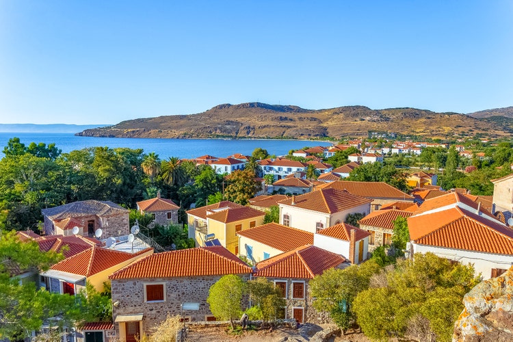 An aerial view of Petra, featuring colorful houses with red roofs surrounded by lush greenery, overlooking sea and hills.
